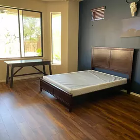 This bright bedroom features warm wood flooring, a dark wooden bed frame with a mattress, and a matching desk by a large window. A dark accent wall with decor adds a modern touch.
