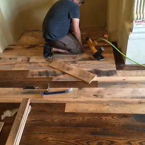 This image captures a person installing mixed hardwood flooring, showcasing various wood tones and textures. Tools like a nail gun and pry bar are visible, indicating an active construction or renovation project.