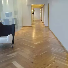 The image features a hallway with a distinctive herringbone pattern hardwood floor, likely showcasing Enmar Hardwood Flooring. The light-filled interior includes a glass-walled office area and white walls, creating a clean, modern aesthetic.