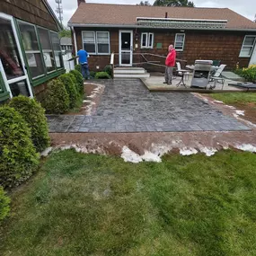 This image features two workers on a backyard patio project. One, in blue, works on a paved section, while another, in red, stands on a wooden deck. The house has a brown exterior and a green sunroom, surrounded by a green lawn.