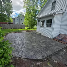This image features a newly installed dark gray paver patio with a curved edge, extending from a white house. The lush green lawn surrounds the new hardscape, and a wooden fence is visible in the background under a cloudy sky.