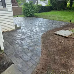 This image features a newly installed dark gray paver patio with a curved edge, extending from a white-sided house into a grassy backyard. Small black landscape lights are visible, and a square stone slab rests on the bare dirt.