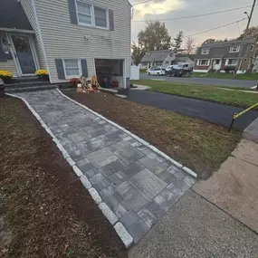 This image shows a newly installed gray paver walkway with granite block edging leading to a light-colored house. A freshly graveled driveway is visible to the right, and the overall scene suggests recent landscaping work.