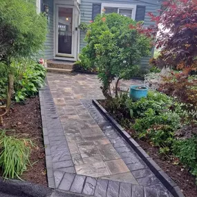 This image features a newly installed multi-toned paver pathway leading to the front door of a light blue house. Darker pavers frame the lighter central path, and lush green landscaping with mature trees surrounds the inviting entrance.