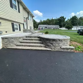 This image features a newly designed outdoor living space with a multi-tiered retaining wall and steps made of stone blocks, leading from a driveway to a house with beige siding. A gravel-filled circular planter adds a decorative touch.