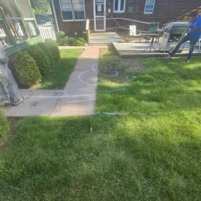 This image captures a backyard in progress, with a long concrete pathway marked with white spray paint for a new design. A worker stands on the left, while a wooden deck and a house with a brown exterior are visible in the background.