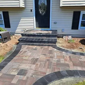 This image showcases a newly installed paver walkway with multi-toned blocks in shades of brown and gray, framed by dark border pavers. It leads to a light-colored house with a dark oval-shaped front door and black shutters.
