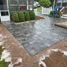 This image features a newly installed dark gray paver patio extending from a house with brown siding and a green sunroom. White sandy residue is visible around the edges, and a worker is on the patio near a white picket fence.