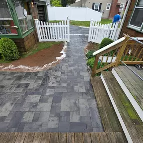 This image shows a newly installed dark gray paver pathway that extends from the edge of a wooden deck. The path is bordered by fresh dirt and leads towards a white picket fence, suggesting recent landscaping work.