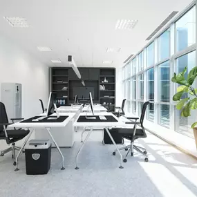 A bright, modern office space features white desks with black computers and chairs, set against a backdrop of large windows offering ample natural light. A green plant adds a touch of life to the contemporary design.