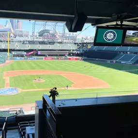 A wide shot of a baseball stadium from a high vantage point, showing the lush green field, empty stands, and a large scoreboard. Two monitors in the upper right display the team logo and a live feed.