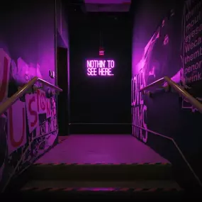 This atmospheric image shows a dimly lit hallway with purple neon lighting, leading down a short flight of stairs. A prominent neon sign reads 