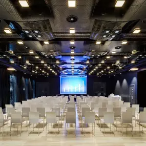 A spacious, modern conference hall is set up for a presentation, featuring rows of white chairs facing a large projection screen displaying a bar graph. The dark ceiling is adorned with numerous lights, creating an immersive ambiance.