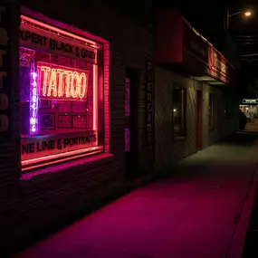 This striking night scene features a tattoo parlor bathed in vibrant pink and purple neon light, illuminating the street and sidewalk. The glow emphasizes 