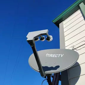 A clear shot of a gray DIRECTV satellite dish mounted on the side of a building with white siding and green trim. The dish is prominently displayed against a vibrant, clear blue sky with power lines.
