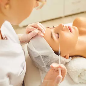 A professional wearing glasses and white gloves is performing a cosmetic injection on a person's upper cheek, near the eye, as the person lies on a white towel-covered bed.