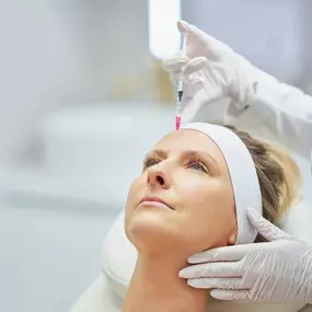 A professional is administering an injection into a woman's forehead as she lies on a white bed with her head elevated. The medical professional wears white gloves and a white coat.