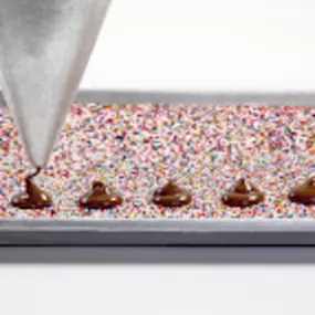 A close-up of bulk chocolate being freshly piped onto a bed of colorful nonpareils. High-quality methods used by wholesale candy suppliers to create gourmet chocolate pieces for retailers and specialty shops.