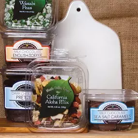 Assorted bulk nuts, dried fruit, and premium snack mixes in clear tubs, including wasabi peas, nut blends, chocolate pretzels, and toffee, ideal for wholesale distributors and specialty retailers.