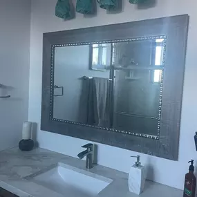 This image showcases a clean and modern bathroom vanity area, featuring a large framed mirror with decorative studs, a white sink set in a light-colored countertop, and a sleek faucet. Above, a light fixture with four teal-tinted glass shades provides illumination.