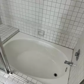 This image showcases a pristine white bathtub with a built-in tiled bench, surrounded by clean white square tiles on the walls and floor. The chrome fixtures gleam, reflecting a meticulously clean and well-maintained bathroom, ready for use.