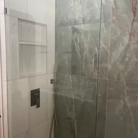 This image features a sparkling clean modern shower with clear glass enclosures and large gray-veined tiles. A square rainfall showerhead and handheld sprayer indicate contemporary design. Recessed shelving is visible, showcasing a well-maintained, minimalist bathroom.
