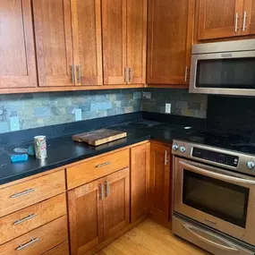 This image shows a kitchen with warm-toned wooden cabinets, dark countertops, and a multi-colored stone tile backsplash. A stainless steel oven and microwave are visible, along with a cutting board and cleaning supplies on the counter, suggesting a pre-cleaning state.