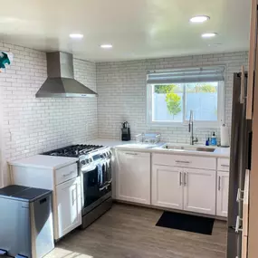 This bright kitchen features white subway tile backsplash, white cabinets, and stainless steel appliances, including a gas range with an overhead range hood. Sunlight streams in through a window above the sink, highlighting the clean, modern space.