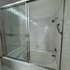 This image displays a clean bathroom with a white tiled shower-tub combo, enclosed by sliding glass doors with chrome frames. A built-in bench, showerhead, and grab bar are visible, all sparkling clean, suggesting a recently serviced bathroom.