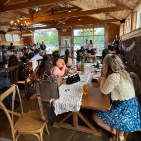 A wide view of a rehearsal dinner hosted at Tranquillity Vineyard & Winery in Middlebury, Connecticut. Guests sit at long wooden tables adorned with greenery and wine, attentively watching a couple near the front of the room.