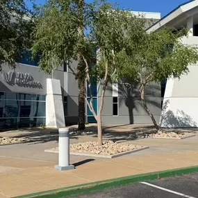 Medical Plaza:   This photo showcases the modern exterior of a professional medical plaza, prominently featuring VIVA MedSuites. The building is architecturally striking with a large arched entryway, clean gray-and-white design, and sleek glass windows along the front. Desert landscaping with palm trees and native greenery lines the sidewalk, while accessible parking is available directly in front of the entrance. The environment reflects a clean, professional, and welcoming setting, ideal for h