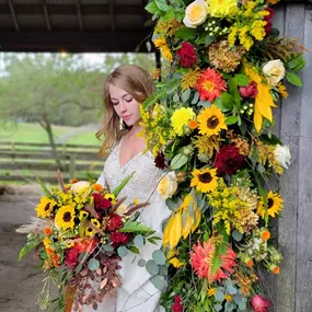 A bride posing by a barn door that is decorated with a large vertical flower arrangement while she is looking down at a bouquet designed by Christina Price of Homestead Floral Wedding & Events.