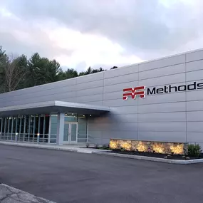 Modern exterior of the Methods Machine Tools headquarters in Acton, Massachusetts, featuring sleek metal siding, large glass windows, and landscaped accents.