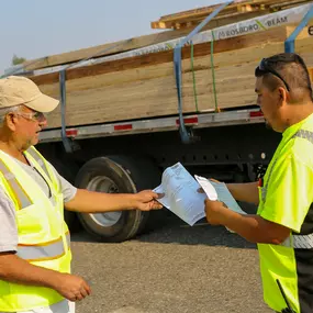 Two Builders FirstSource employees wearing high-visibility safety vests are reviewing paperwork in front of a flatbed truck loaded with lumber. One worker is handing documents to the other as they stand in a paved outdoor yard, ensuring proper delivery and coordination of building materials.