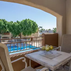A table with a bowl of fruit on it sits on a balcony overlooking a pool.