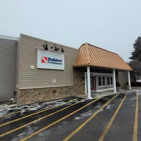 A photo of the Builders FirstSource storefront in Auburn Hills, MI, during winter. The building features light beige siding with a stone facade along the lower portion and a copper-colored metal awning supported by white pillars above the entrance. The company’s logo is prominently displayed on a white sign with black overhead lighting fixtures. The parking lot in the foreground shows snow lightly scattered across the asphalt, with yellow parking lines visible. Leafless trees and evergreen shrub