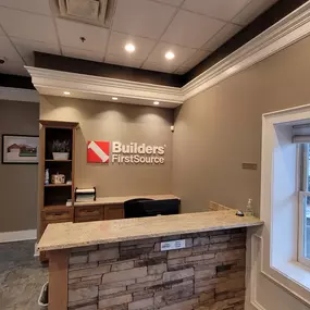 A welcoming interior view of the Builders FirstSource front desk at the Auburn Hills, MI location. The reception area features a beige wall with the company logo displayed prominently in white and red lettering under warm recessed lighting. The desk has a stone facade and a polished granite countertop with a small bell and hand sanitizer placed on top. A wooden shelving unit to the left holds decorative items and brochures, while a window to the right provides a view of the parking lot and shrub