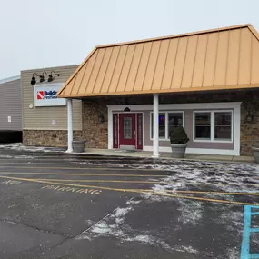 The exterior view of the Builders FirstSource location in Auburn Hills, MI, showcasing the front entrance and parking lot on a winter day. The building features beige siding and a stone facade with a copper-colored metal awning supported by white columns. The red front doors are framed by large white-trimmed windows, with decorative planters placed on both sides. Above the entrance, the Builders FirstSource logo is prominently displayed under black light fixtures. The parking lot is lightly dust
