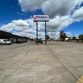 A large parking lot in front of a Builders FirstSource building. The prominent company sign is displayed on a tall pole in the center of the image, with the building on the left side and some parked vehicles, including a white service van. The parking lot is mostly empty, and the sky is filled with puffy clouds against a bright blue background. Sparse trees and bushes line the parking lot.