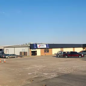 A wide-angle view of a Builders FirstSource facility, including a parking lot and storefront. The building features a mix of brick and metal siding with large signage displaying the Builders FirstSource logo above the entrance and on a standalone sign to the right. Several vehicles, including trucks and a red dump truck, are parked in the lot. The facility appears to include multiple sections, with a lumber yard visible in the background. The sky is clear and blue, and the overall scene reflects