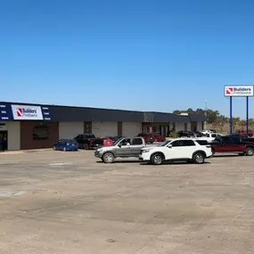 A wide shot of a Builders FirstSource facility with a parking lot in the foreground. The building has a mix of brick and metal siding with large signage displaying the Builders FirstSource logo above the entrance. Several cars and trucks are parked in the lot. To the right, a large standalone sign with the Builders FirstSource logo is visible near the street. The surrounding area includes trees and other commercial buildings under a clear blue sky, indicating a sunny day.