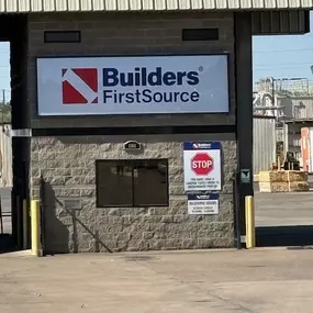 A close-up view of a Builders FirstSource entrance gate and receiving area. The sign with the Builders FirstSource logo is prominently displayed above a small window in a stone-clad structure. Below the sign, there are additional instructions, including a red stop sign that reads 