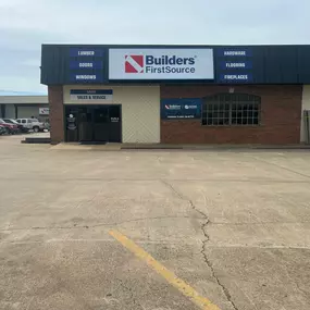 The front of a Builders FirstSource building featuring a large sign with the company logo and a list of services, including lumber, doors, windows, hardware, flooring, and fireplaces. The building has a mix of brick and light-colored siding, with a glass door marked 