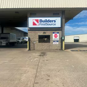 The entrance to the receiving area at a Builders FirstSource facility. A large sign with the company logo is displayed above a small window on a stone-clad building. A 