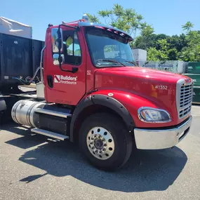 Builders FirstSource flatbed delivery truck with red cab.