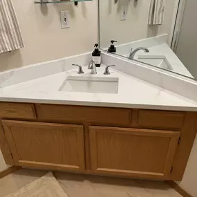 A modern bathroom vanity with a white countertop and sink, light wood cabinets, and a mirror reflecting the room.