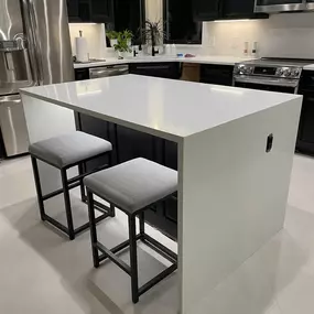 A modern kitchen with a large white island, two gray bar stools, black cabinets, and stainless steel appliances.
