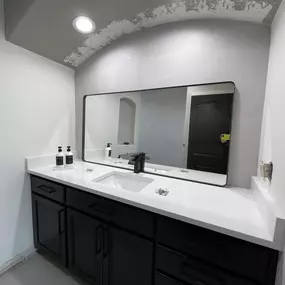 A modern bathroom vanity with a white countertop, black cabinets, a large mirror, and two soap dispensers.