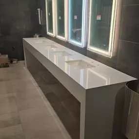 A modern public restroom with a long white countertop featuring multiple sinks and three illuminated mirrors.