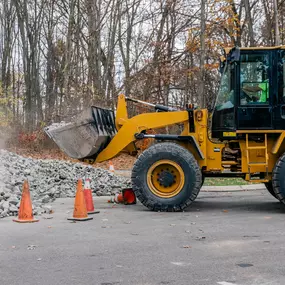 Yellow front loader scooping gravel surrounded by orange cones in a wooded area.